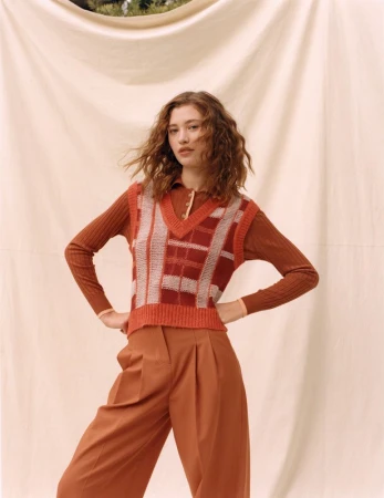 A young woman poses in an outfit with a brown color palette, wearing a knit sweater vest and culottes against a draped backdrop.