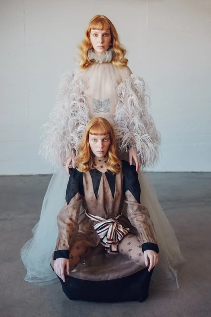 Two red-haired women in artistic gowns pose in a studio, one seated and one standing behind her, captured in a fashion-forward style.