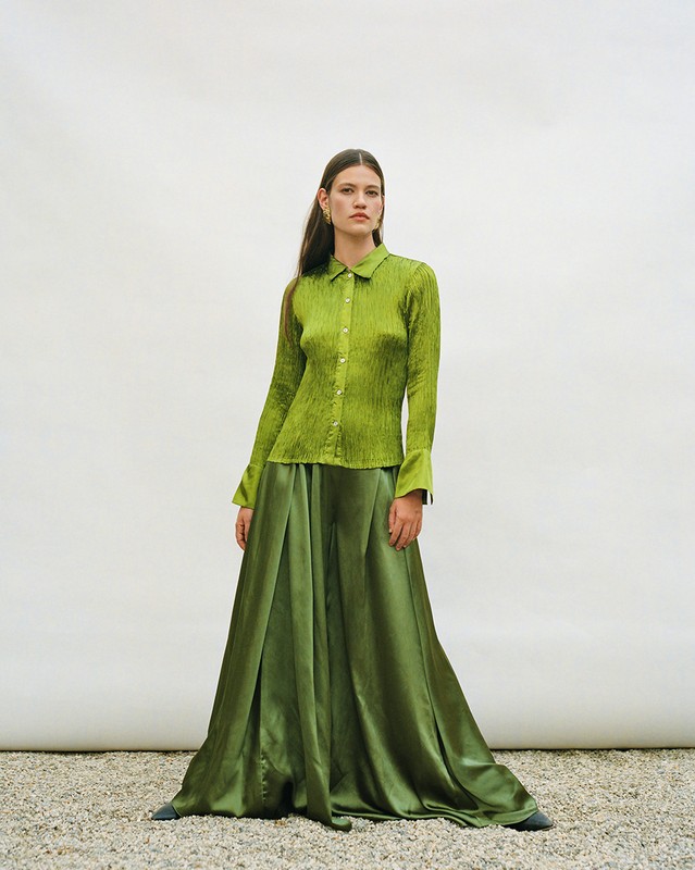 Full-length shot of a woman in green satin blouse and skirt against a pale backdrop with gravel ground. Fashion editorial.