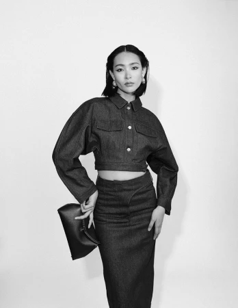Black and white portrait of a woman wearing a cropped jacket and skirt holding a clutch against a white seamless background.