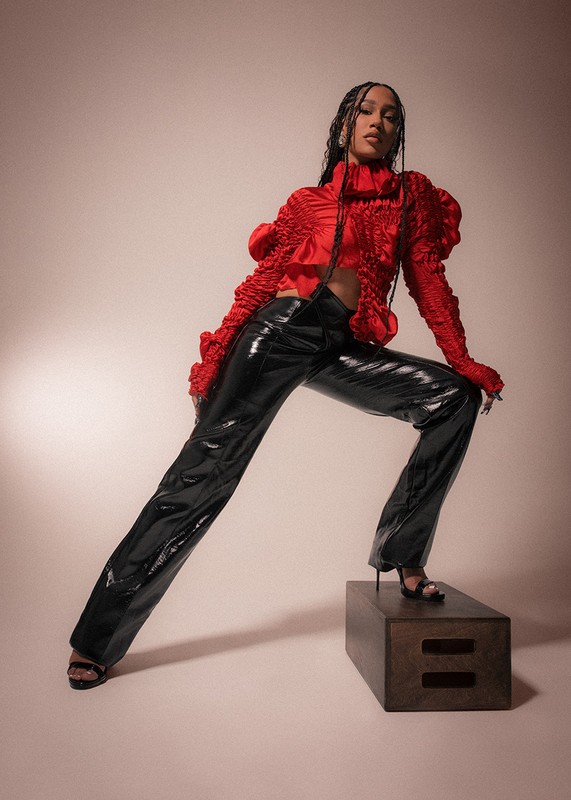Stylish woman poses on a box in red top with ruffled sleeves and black shiny pants in a studio. Beauty and fashion photography.