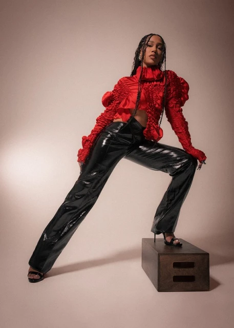 Stylish woman poses on a box in red top with ruffled sleeves and black shiny pants in a studio. Beauty and fashion photography.