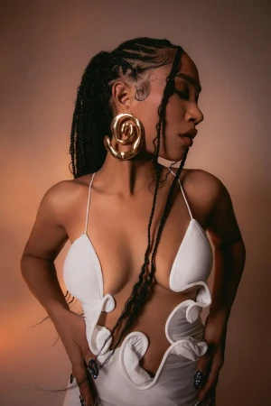 Close-up portrait of a Black woman wearing a white cutout dress and large gold spiral earrings with braided hair.