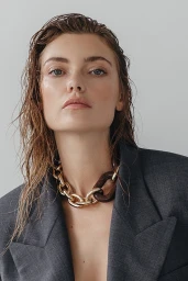 Close-up portrait of a woman with wet hair wearing a grey blazer and a chunky gold and wood link necklace