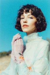 Woman with short dark curly hair wearing sheer pale top and gloves against bright blue sky background