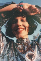 Close-up portrait of a smiling woman in a shiny silver sequined top with her hands raised to her head, wearing a chunky gemstone bracelet.