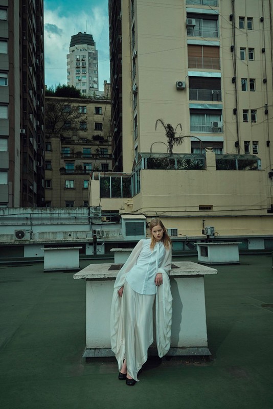 Model wearing a white blouse and long skirt leaning against a rooftop structure framed by tall urban apartment buildings.