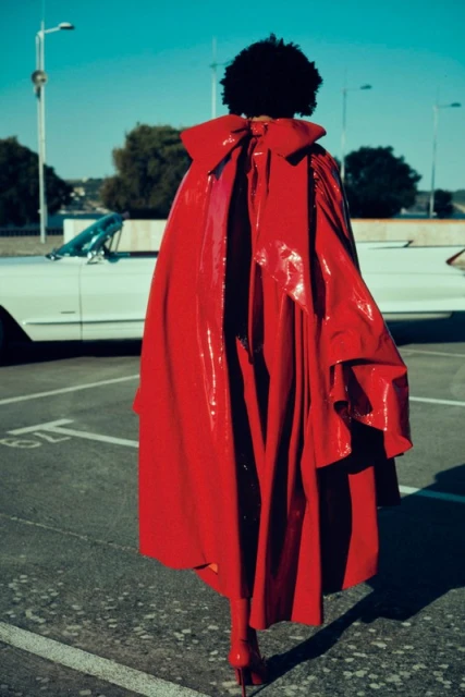 A woman in a red patent leather cape with a large bow poses in a parking lot, standing near a vintage white car under a clear sky.