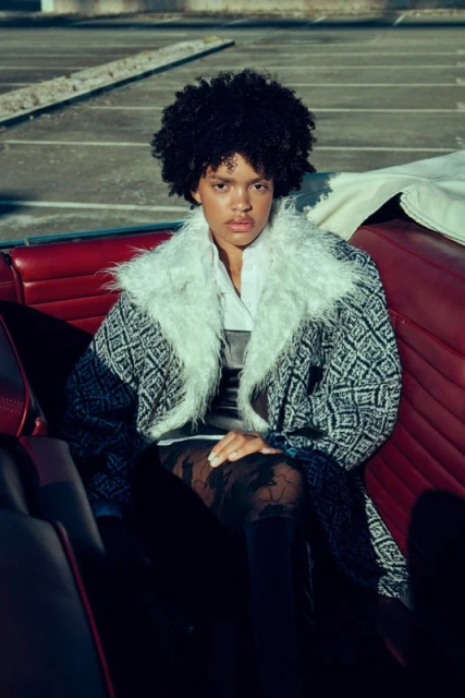 Stylish woman with afro seated in vintage red convertible, wearing patterned coat with fur collar, black leggings, posing in parking lot.