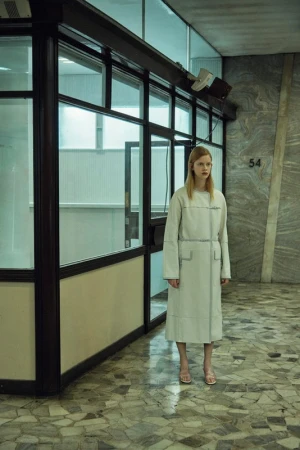 Model in a cream white coat standing in a marble hallway by glass walls photographed by Sofia and Mauro.