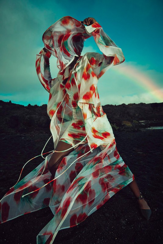A fashionable woman in a translucent, watercolor-patterned dress poses in a lava field under a rainbow sky, capturing a blend of fashion and nature.