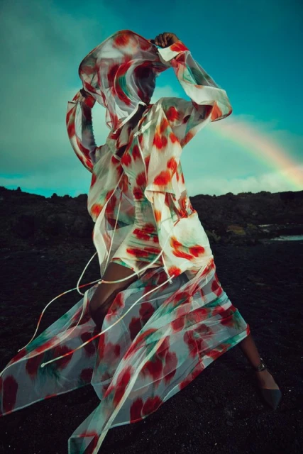 A fashionable woman in a translucent, watercolor-patterned dress poses in a lava field under a rainbow sky, capturing a blend of fashion and nature.