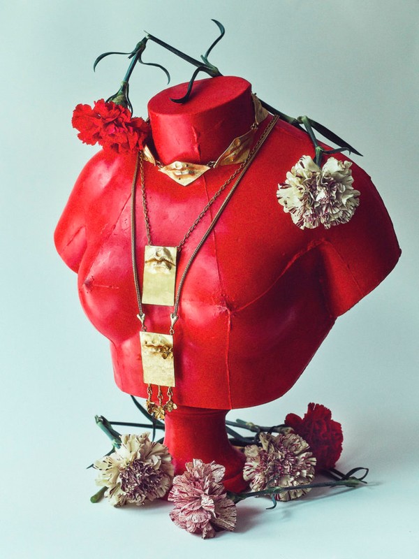 Red mannequin bust with gold jewelry and carnation flowers in a studio setting, showcasing fashion and artistic still life.