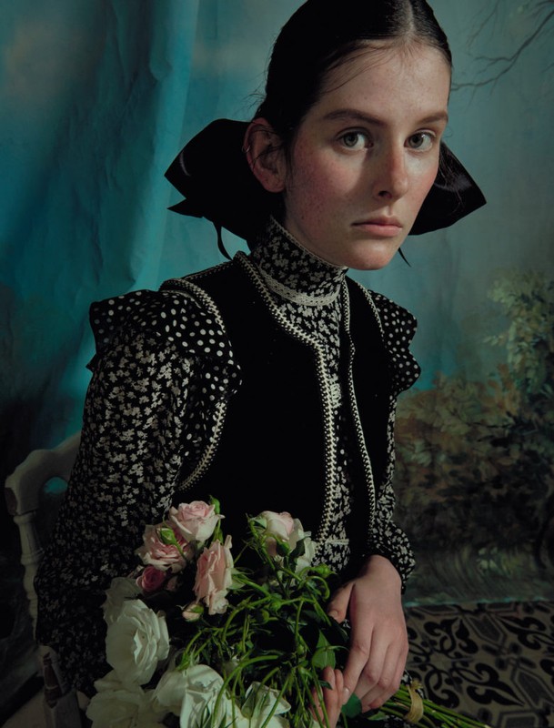 Portrait of a young woman with fair skin and dark hair in a patterned black dress holding a bouquet of roses against a teal backdrop.