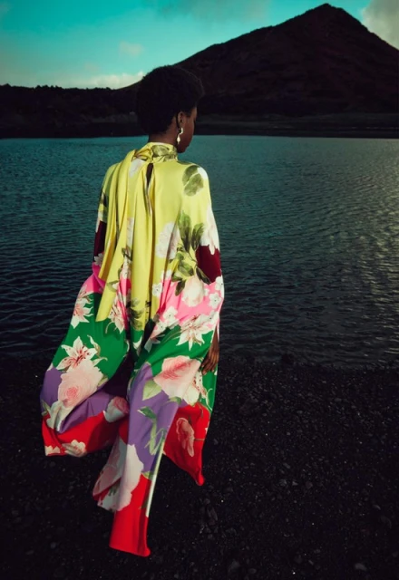 Woman in floral print dress stands near dark lake with mountain backdrop under teal sky, captured at eye-level in outdoor setting.