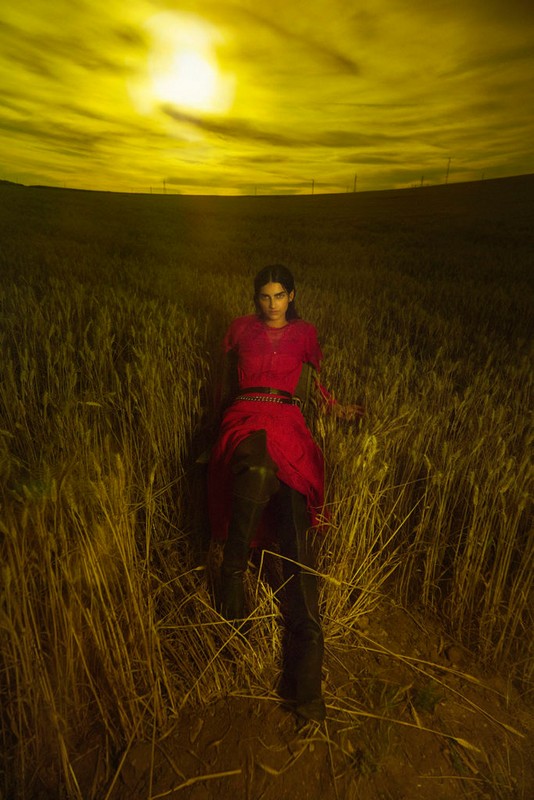 A woman in a red dress stands in a wheat field under a bright yellow sky, creating a surreal and eye-catching fashion portrait.