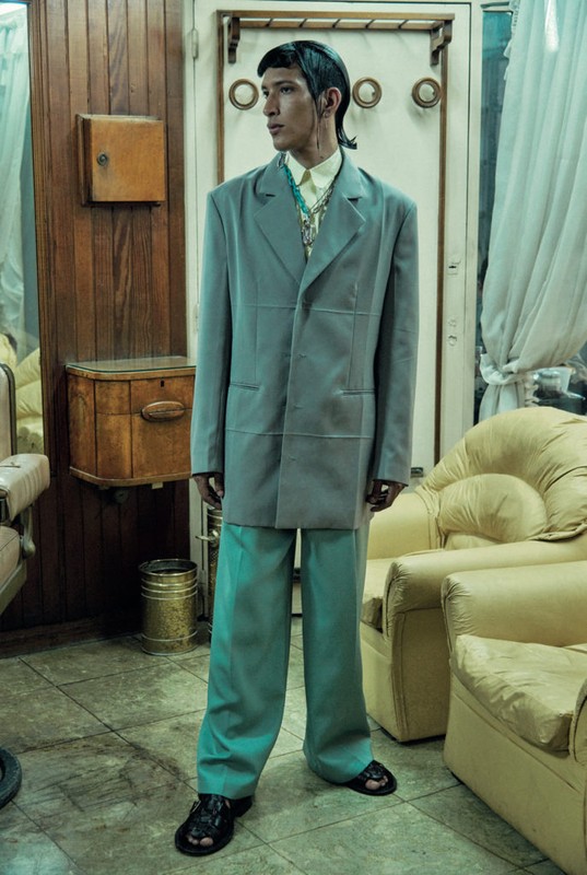Young man in oversized teal suit in vintage barber shop with aged wood paneling, cream chairs, mirrors, and aged tile floor.