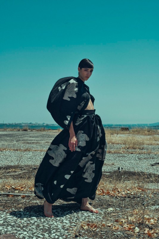 An adult woman models a voluminous, patterned black skirt and cropped top against an arid landscape under a clear blue sky.