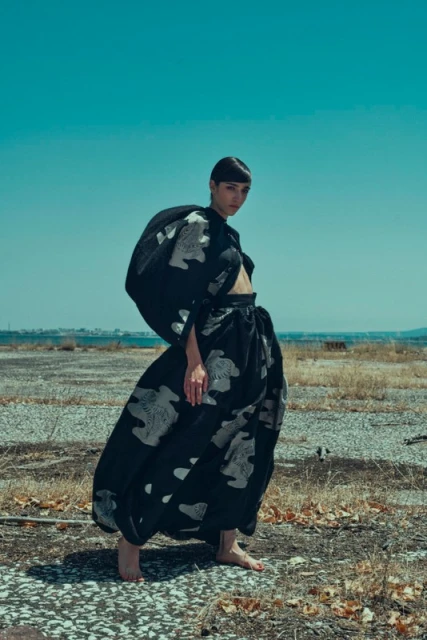 An adult woman models a voluminous, patterned black skirt and cropped top against an arid landscape under a clear blue sky.