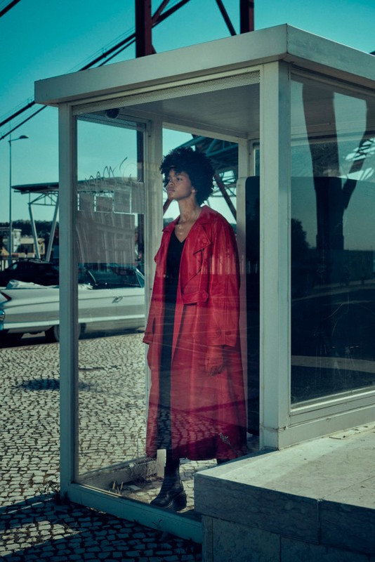 Woman in a red raincoat stands inside a glass bus stop against a backdrop of a bridge, sky and street with a car on a sunny day.