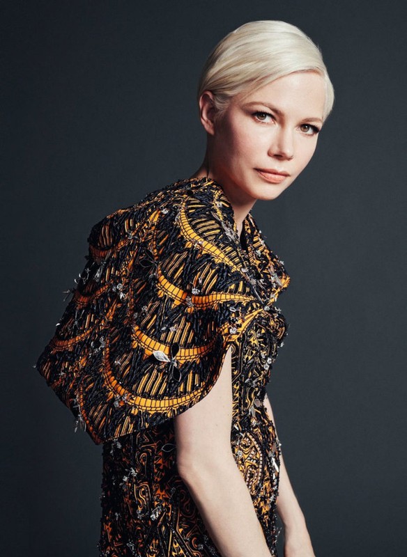 Elegant studio portrait of fair-skinned adult woman with blonde pixie cut, wearing black and gold patterned cape dress.