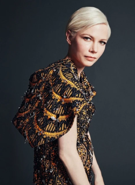 Elegant studio portrait of fair-skinned adult woman with blonde pixie cut, wearing black and gold patterned cape dress. 
