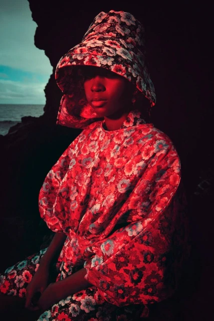 Woman in a floral dress and hat sits near a rock formation against a moody ocean backdrop, creating a fashion editorial scene.