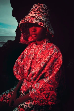Woman in a floral dress and hat sits near a rock formation against a moody ocean backdrop, creating a fashion editorial scene.