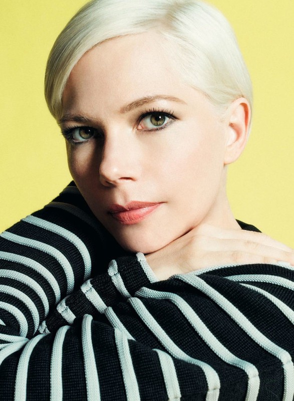 Close-up portrait of a woman with short platinum blonde hair wearing a black and white striped sweater against a yellow background.