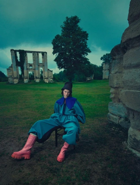 Woman in blue jumpsuit and pink boots sits in grassy field with crumbling ruins and a cloudy sky, creating a striking outdoor fashion editorial.