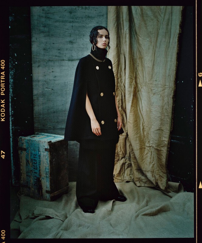 A stylish fashion shot features a model in an elegant black cape and wide-leg pants, posing in front of a rustic curtain and antique box.