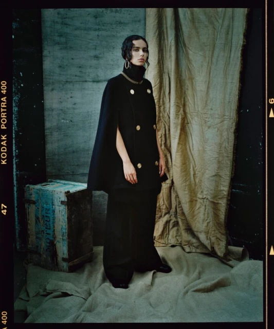 A stylish fashion shot features a model in an elegant black cape and wide-leg pants, posing in front of a rustic curtain and antique box.