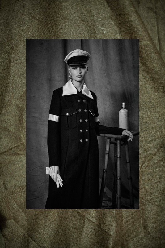 Black and white portrait of a woman in a dark coat, cap, and gloves, standing in front of a neutral backdrop with a stool and bottle.