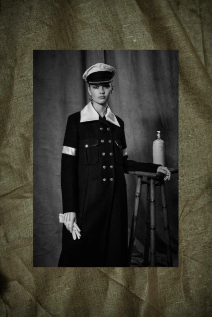 Black and white portrait of a woman in a dark coat, cap, and gloves, standing in front of a neutral backdrop with a stool and bottle.