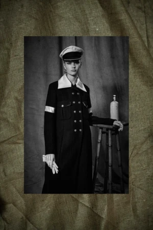 Black and white portrait of a woman in a dark coat, cap, and gloves, standing in front of a neutral backdrop with a stool and bottle.
