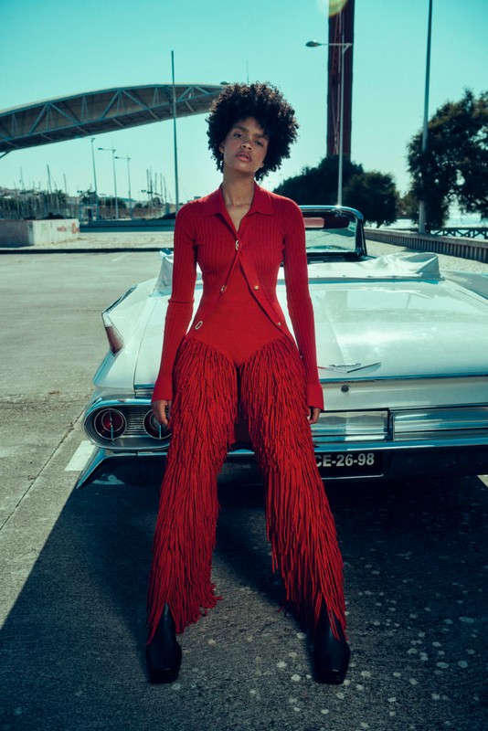 A stylish adult female with afro hair poses in red fringe outfit next to a white vintage car on a sunny city street with a bridge.