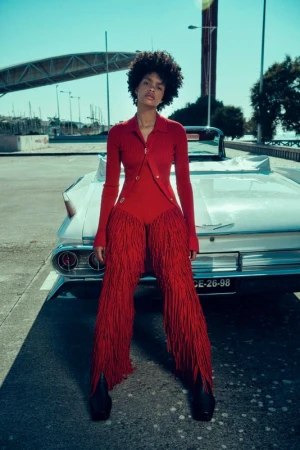 A stylish adult female with afro hair poses in red fringe outfit next to a white vintage car on a sunny city street with a bridge.