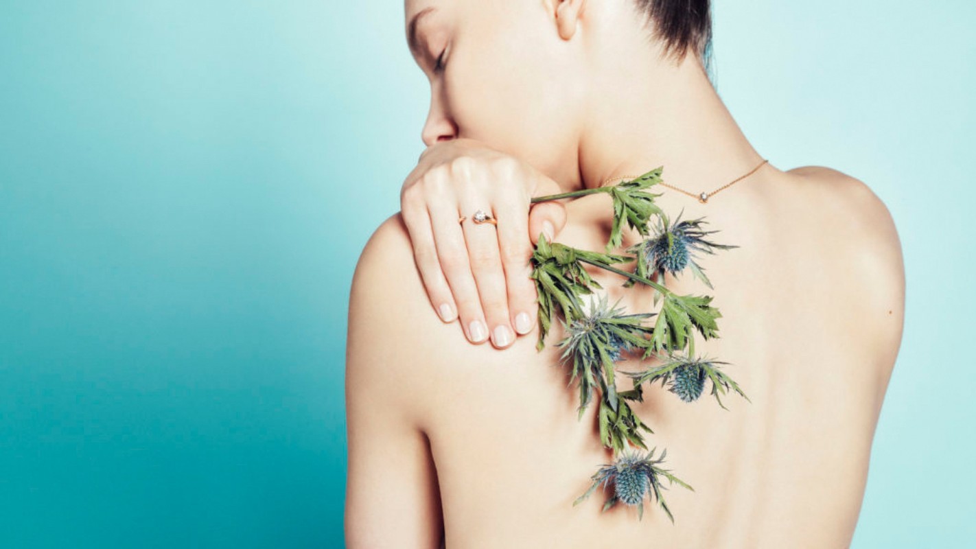 Close-up of a woman's bare back and shoulder with her hand and a ring holding a sprig of thistle against a light blue background.
