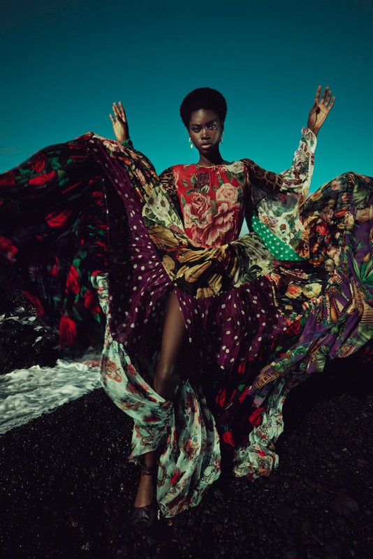 A striking image showcases a Black female model in a flowing, multicolored dress on a rocky beach with a turquoise sky backdrop.