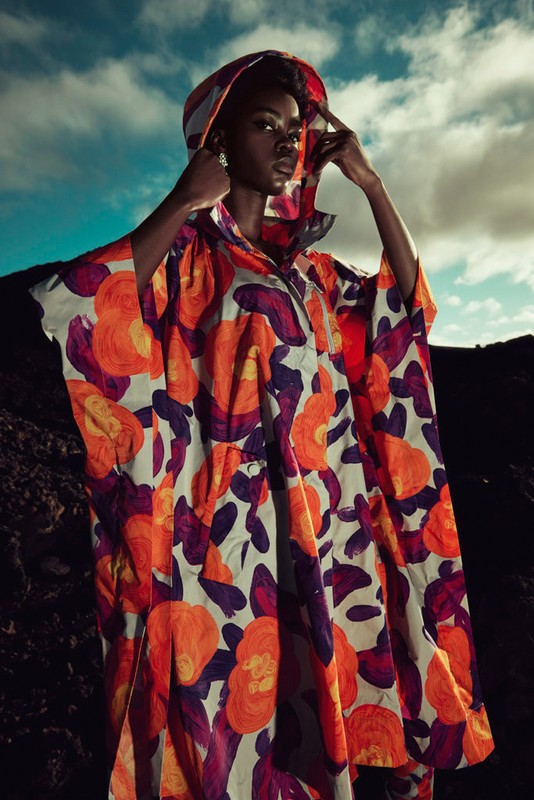 Woman poses wearing floral hooded cape in outdoor setting, striking a pose with hands near her head, set against a cloudy sky.