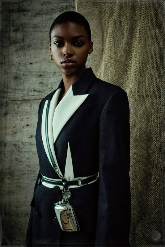 A stylish black woman with short hair poses in a black blazer with white trim and a silver pendant in front of a textured backdrop.