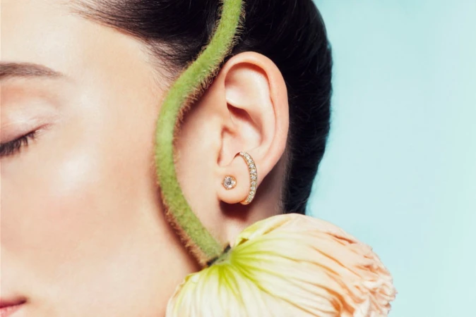 Close-up portrait of a woman wearing diamond earrings with a green stem and pink flower curving around her ear against a pale blue background.