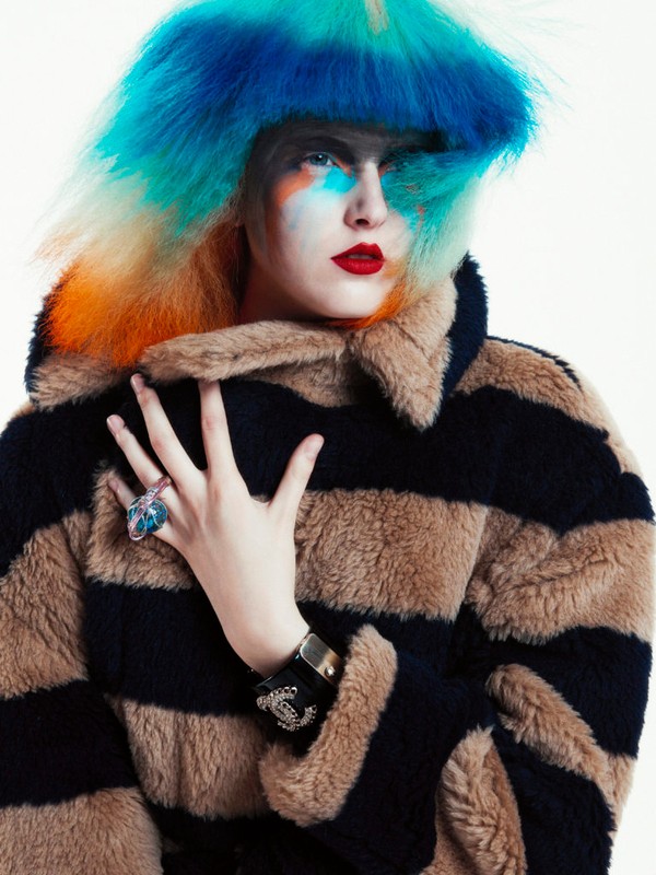 Fashion shot of a woman with bright blue, orange, and blonde hair wearing a striped faux fur coat against a bright white backdrop.