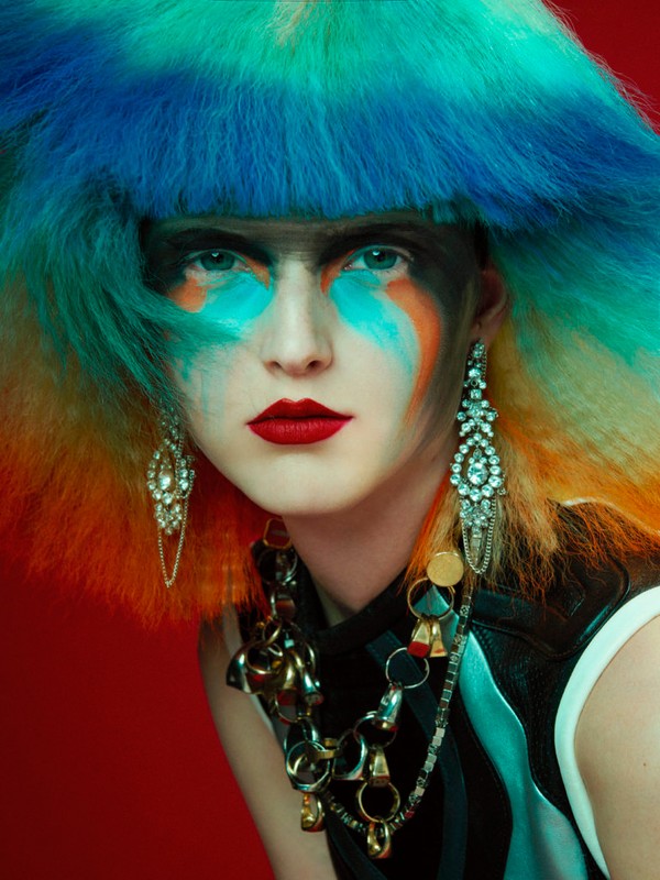 Eye-level portrait of a woman with bright makeup and a colorful wig against a red background, focusing on her face, jewelry, and dramatic style.