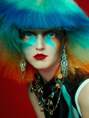 Eye-level portrait of a woman with bright makeup and a colorful wig against a red background, focusing on her face, jewelry, and dramatic style.