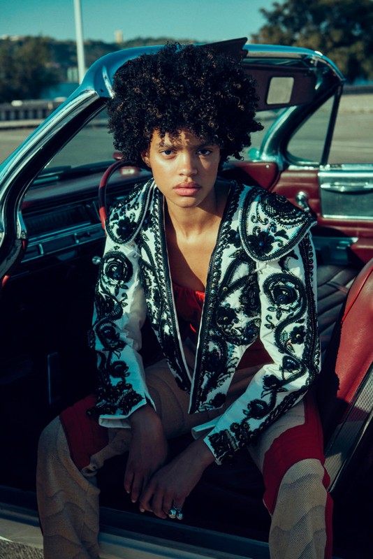 A young woman with curly hair in an embroidered jacket sits inside a vintage convertible car on a bright day, looking at the camera.