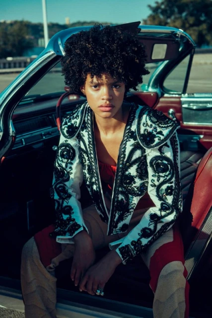 A young woman with curly hair in an embroidered jacket sits inside a vintage convertible car on a bright day, looking at the camera.