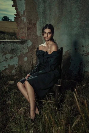 Woman in dark off-shoulder dress seated on a wooden chair amidst overgrown grass before a weathered wall by Sofia & Mauro