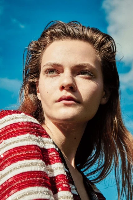 Portrait of a woman with light brown hair, wearing a red and white striped top, against a blue sky with scattered clouds.