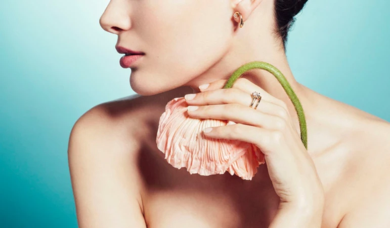 Close-up portrait of a woman holding a pale pink flower against her chin against a soft blue gradient background.